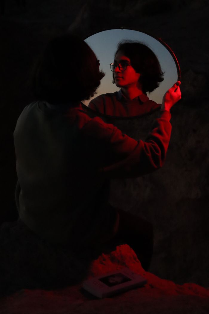 A woman in red light reflects in a mirror in a dark environment, creating a moody atmosphere.